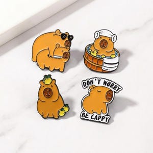 Puede incluir: Cuatro pines de esmalte con capibaras en varias poses. Uno lleva gafas de sol, otro se relaja en una bañera de madera, un tercero tiene fruta en la cabeza y el último dice "Don't Worry Be Cappy".