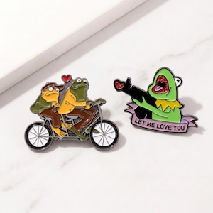 Könnte beinhalten: Zwei Emaille-Anstecknadeln mit Froschfiguren. Eine Nadel zeigt zwei Frösche, die Fahrrad fahren, mit einem Herz darüber. Die andere Nadel zeigt einen grünen Frosch, der eine Waffe hält, mit dem Text "LET ME LOVE YOU" auf einem Banner.