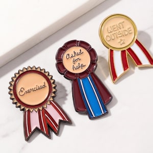 May include: Three enamel pins in the shape of ribbons. One pin says "Exercised" with pink and red ribbons. Another says "Asked for help" with blue and burgundy ribbons. The third says "Went Outside" with red and white ribbons.