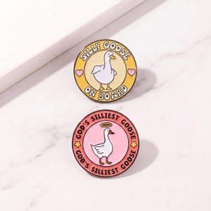 May include: Two circular enamel pins. The top pin has a yellow border, a white goose, and the text SILLY GOOSE ON BOARD with two pink hearts. The bottom pin has a pink centre, a white goose with a halo, and the text GOD'S SILLIEST GOOSE with two yellow stars.