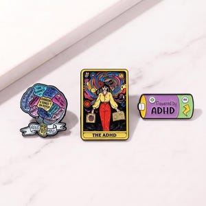 May include: Three enamel pins: a brain-shaped pin labeled ADHD MIND with text segments, a rectangular tarot-style pin titled THE ADHD featuring a person surrounded by colorful swirls, and a purple battery-shaped pin labeled Powered by ADHD.