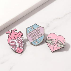 Könnte beinhalten: Drei Emaille-Anstecknadeln: ein rosa anatomisches Herz mit Brustkorb, ein Schild mit dem Text "Pointless Meeting Survivor" und ein rosa Herz mit Skeletthänden. Die Anstecknadeln haben schwarze Umrisse.