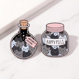 Op de afbeelding: Twee emaille pins: een zwarte fles met een kurk en de tekst "Happiness Potion" op een label, gevuld met grijze en zwarte kattengezichten. De andere is een pot "Happy Pills" met grijze en zwarte kattengezichten.
