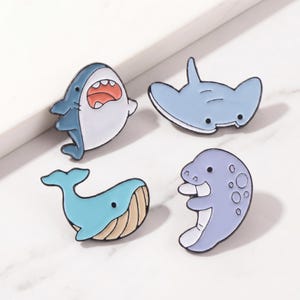 Puede incluir: Conjunto de cuatro broches de esmalte con diseños de vida marina. Incluye un tiburón azul y blanco, una manta raya azul claro, una ballena azul y un manatí morado. Cada broche tiene un contorno negro.