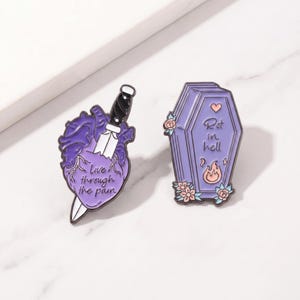 May include: Two enamel pins. One is a purple heart pierced by a knife, with the text "Live through the pain." The other is a purple coffin with the text "Rot in hell" and a flame graphic.