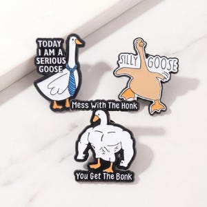 Pode incluir: Três pins de esmalte com desenhos de gansos de desenho animado. Um mostra um ganso com gravata e o texto "TODAY I AM A SERIOUS GOOSE". Outro pin apresenta um ganso com o texto "SILLY GOOSE". O terceiro pin mostra um ganso musculoso com o texto "YOU GET THE BONK".