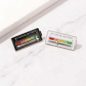 May include: Two enamel pins with a battery gauge design. One pin is black with a white gauge, and the other is white with a black gauge. Both pins read "MY SOCIAL BATTERY" and have a sliding indicator.