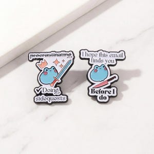 Puede incluir: Dos pines esmaltados con un diseño de rana. Un pin dice "procrastinating" y "Doing sidequests" con una rana sosteniendo una espada. El otro pin dice "I hope this email finds you" y "Before I do" con una rana sosteniendo un cuchillo.