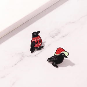 Puede incluir: Dos pines de esmalte con forma de cuervos negros. Uno lleva un suéter rojo con un patrón geométrico blanco y pequeñas siluetas de pájaros negros. El otro cuervo lleva un sombrero rojo con lunares blancos y un ala de color verde claro.