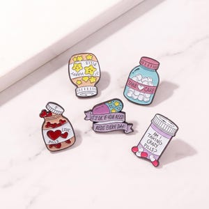 Könnte beinhalten: Fünf Emaille-Anstecknadeln mit medizinischen Motiven: ein Glas mit gelben sternförmigen Pillen mit der Aufschrift HAPPY PILLS, eine blaue Flasche mit TAKE CARE, ein Glas mit roten herzförmigen Pillen mit LOVE PILLS, eine Kapsel auf einem Banner mit IT'S OK IF YOU NEED MEDS EVERY DAY und eine Flasche mit AM I TAKING CRAZY PILLS?