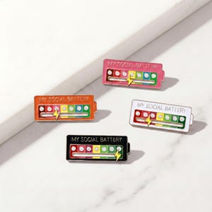 May include: Four enamel pins in pink, orange, white, and black, each with the text "MY SOCIAL BATTERY." The pins feature a battery-like design with a slider indicating social energy levels, from red to green, and a lightning bolt.