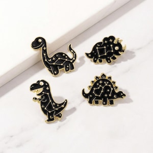 May include: Four black dinosaur pins with gold-colored outlines. Each pin features a different dinosaur shape, with a constellation design in white. The pins are set against a white background.