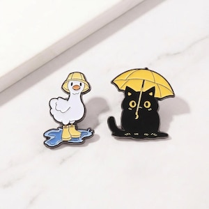 May include: Two enamel pins: a white duck wearing a yellow hat and boots standing in a blue puddle, and a black cat under a yellow umbrella. The pins have black outlines and are on a white background.