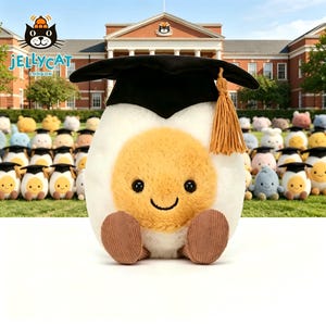 May include: A plush, smiling egg character wearing a black graduation cap with a gold tassel. The egg has a yellow yolk and white egg white. Other plush eggs wearing graduation caps are in the background. The Jellycat London logo is in the upper left corner.