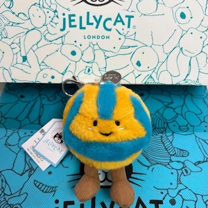 May include: A plush, round Jellycat keychain in yellow and blue, resembling a volleyball, with brown corduroy legs. The keychain has a smiling face and is displayed on a box with the Jellycat logo and illustrations.