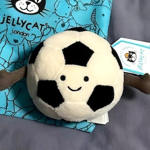 May include: A plush soccer ball toy with a smiling face and brown stick-like arms. The ball is white with black pentagon shapes. The toy is from Jellycat London, with a tag that reads "Jellycat Sports". The background includes a blue bag with a repeating pattern.