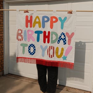 May include: A white fabric wall hanging banner featuring the colorful appliqued text HAPPY BIRTHDAY TO YOU in various patterns and colors. The banner is suspended from a wooden rod and finished with a red fringe trim along the bottom edge.