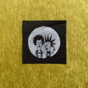 May include: A square black fabric patch featuring a circular white graphic print of two cartoon characters. One character has messy dark hair, and the other has a tall spiked mohawk. Both figures are wearing t-shirts with simple graphic designs.