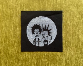 May include: A square black fabric patch featuring a circular white graphic print of two cartoon characters. One character has messy dark hair, and the other has a tall spiked mohawk. Both figures are wearing t-shirts with simple graphic designs.