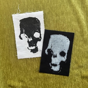 May include: Two rectangular fabric patches featuring skull illustrations. One patch is white with a black ink skull print, and the other is black with a white ink skull print. Both patches have raw, frayed edges and a high-contrast graphic design.