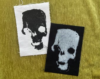 May include: Two rectangular fabric patches featuring skull illustrations. One patch is white with a black ink skull print, and the other is black with a white ink skull print. Both patches have raw, frayed edges and a high-contrast graphic design.
