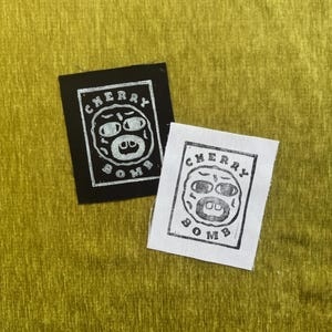 May include: Two square fabric patches featuring a graphic print of a face inside a rectangular border with the text CHERRY BOMB. One patch is black with a white print, and the other is white with a black print, both displaying a distressed ink texture.