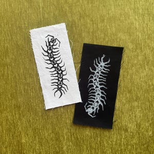 May include: Two rectangular fabric patches featuring a centipede illustration with heart-shaped segments. One patch is white with a black ink print, and the other is black with a white ink print. Both patches have raw, frayed edges on the sides.