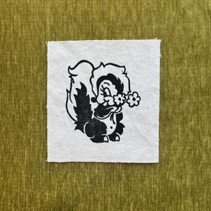 May include: A square white fabric patch featuring a black screen-printed illustration of a cartoon skunk holding a small flower. The patch is centered on a textured olive green fabric background with a subtle vertical grain and soft appearance.