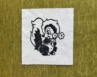 May include: A square white fabric patch featuring a black screen-printed illustration of a cartoon skunk holding a small flower. The patch is centered on a textured olive green fabric background with a subtle vertical grain and soft appearance.