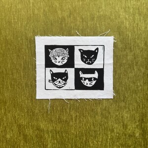 May include: A rectangular white fabric patch featuring a black ink block print of four distinct cat faces arranged in a two-by-two grid. Each cat has a unique expression or accessory, including a beanie, a bandana, and sunglasses, all on a textured olive green background.