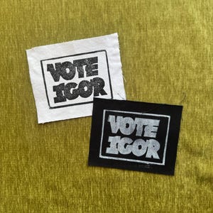 May include: Two rectangular fabric patches featuring the text VOTE IGOR enclosed in a rectangular border. One patch is white with black ink, and the other is black with white ink. Both patches have raw, unfinished edges and a visible woven texture.