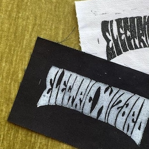 May include: Two rectangular fabric patches, one black with white ink and one white with black ink. Both patches feature the stylized text "ELECTRIC WIZARD" printed in a bold, distorted font. The edges of the fabric are raw and slightly frayed.