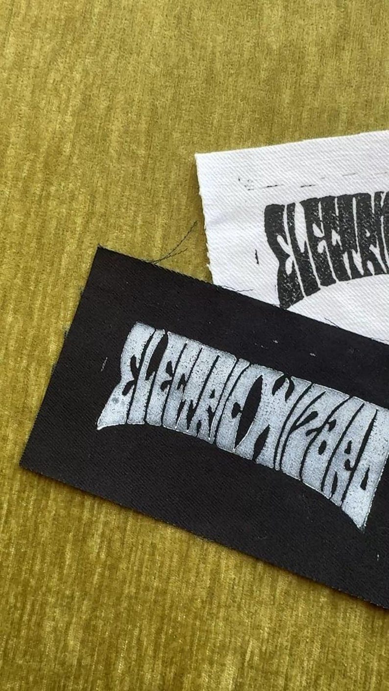May include: Two rectangular fabric patches, one black with white ink and one white with black ink. Both patches feature the stylized text "ELECTRIC WIZARD" printed in a bold, distorted font. The edges of the fabric are raw and slightly frayed.