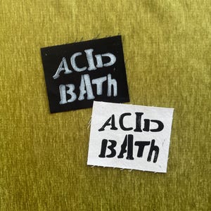 May include: Two rectangular fabric patches featuring the text ACID BATH in a stylized font. One patch is black with white lettering, and the other is white with black lettering. Both patches have raw, slightly frayed edges on all four sides.