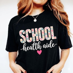 Puede incluir: Camiseta negra con las palabras "SCHOOL" y "health aide". La palabra "SCHOOL" está en letras grandes y florales. El texto "health aide" está en cursiva, con un corazón rosa debajo.