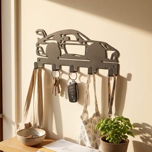 May include: A dark gray metal key holder shaped like a car. The key holder has five hooks and is mounted on a beige wall. Keys, a car key fob, and two tote bags hang from the hooks. A small bowl, a plant, and papers are on a wooden surface below.