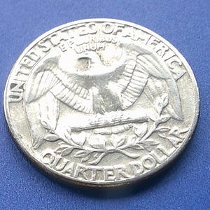 May include: A silver-colored United States quarter dollar coin featuring an eagle with outstretched wings, clutching arrows and an olive branch. The text UNITED STATES OF AMERICA, E PLURIBUS UNUM, and QUARTER DOLLAR are embossed on the coin surface.