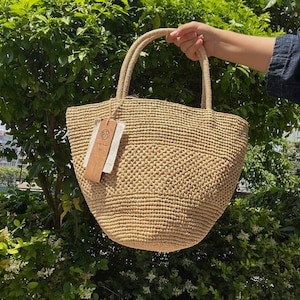 Può includere: Una borsa tote in paglia intrecciata color beige con due manici arrotondati e un'etichetta rettangolare in carta marrone chiaro attaccata sul lato. La borsa presenta un motivo a maglia denso e testurizzato, creando una forma strutturata e flessibile.