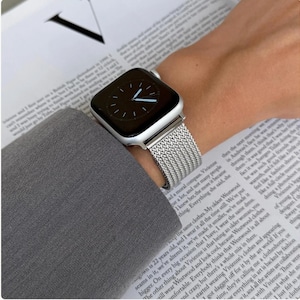 May include: Silver smartwatch with a mesh strap, showing the time. The watch is worn on a wrist, with a grey sleeve in the background. The watch face is black with white hands.