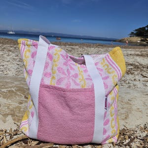 Pode incluir: Uma bolsa de praia rosa e amarela estampada, com alças brancas e um bolso frontal rosa. A bolsa é feita de um material semelhante a uma toalha e está em uma praia arenosa, com o mar ao fundo.