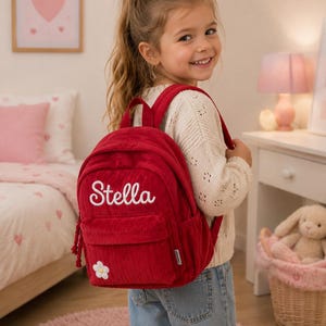 Op de afbeelding: Een dieprode rugzak van corduroy met de naam Stella in wit cursief schrift geborduurd op het voorvak. De tas is voorzien van een kleine witte bloemapplicatie met een geel hartje in de onderste hoek en bijpassende rode schouderbanden.