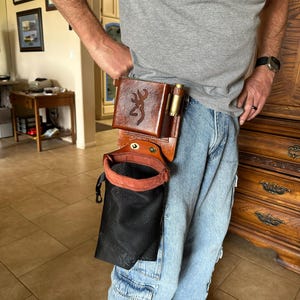 May include: A brown leather shell pouch attached to a belt, featuring a stamped Browning buckmark logo on the front pocket and a side loop holding a brass shotgun shell. Below the leather pocket is a black mesh bag with a brown leather rim.