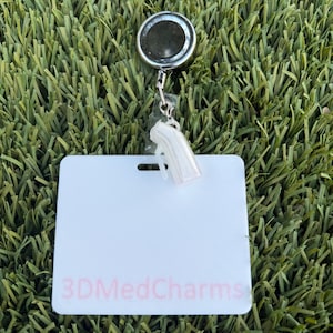May include: A retractable badge reel with a metallic circular housing and a clear plastic strap. Attached to the strap is a small, white, pearlescent hearing aid charm. Below the reel is a white rectangular badge holder with 3DMedCharms printed in pink.