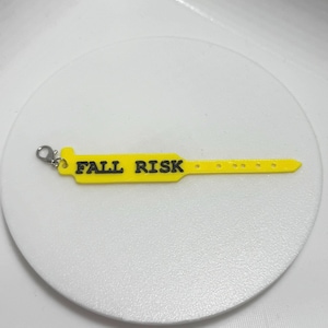 Puede incluir: Una pulsera de plástico amarilla con el texto "FALL RISK" en negro. La pulsera tiene un cierre en un extremo y una serie de agujeros para ajustar. La pulsera está sobre una superficie blanca.
