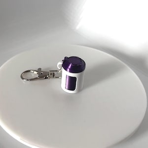 May include: A small, white and purple keychain accessory with a silver clasp. The keychain has a cylindrical shape with a purple top and a white body. The clasp is attached to a silver ring. The item is sitting on a white surface.