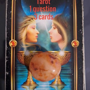 May include: Tarot card with the text "Tarot 1 question 3 cards". The card features two women facing each other, with two golden chalices and a large, round, brown stone. The card reads "TWO OF CUPS".