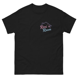 Può includere: T-shirt nero con un piccolo logo colorato sulla parte superiore sinistra del petto. Il logo presenta il contorno di una casa con le parole "Rori's Room" in rosa e blu. La maglietta è realizzata in materiale morbido e ha un classico girocollo.