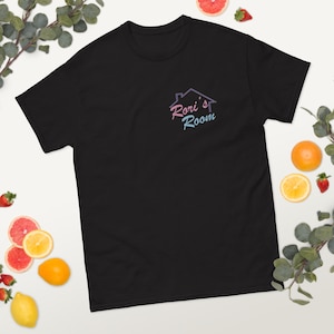 Può includere: T-shirt nero con la scritta "Rori's Room" in un gradiente rosa e blu, con una grafica di una casa. La maglietta è a girocollo.