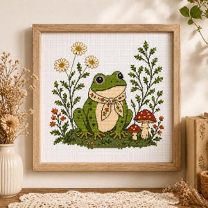 Puede incluir: Arte de punto de cruz de una rana verde con una bufanda con detalles florales, rodeada de flores y setas. La obra está enmarcada en un marco de madera clara. La rana está sentada sobre un parche de hierba verde.