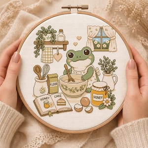 May include: A cross-stitch embroidery hoop featuring a frog chef in an apron, mixing ingredients in a bowl. The scene includes a cookbook, honey jar, flour, and a window with curtains. The color palette is soft, with greens, yellows, and creams.